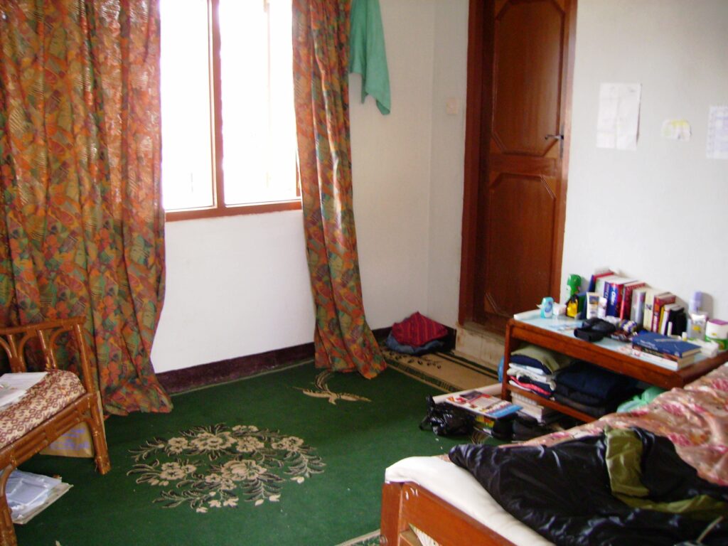 My small room in Kathmandu, with bright curtains framing the sun coming through the window. There's a bed, a chair, a small piece of furniture with books and sunscreen and a door leading to the main house.